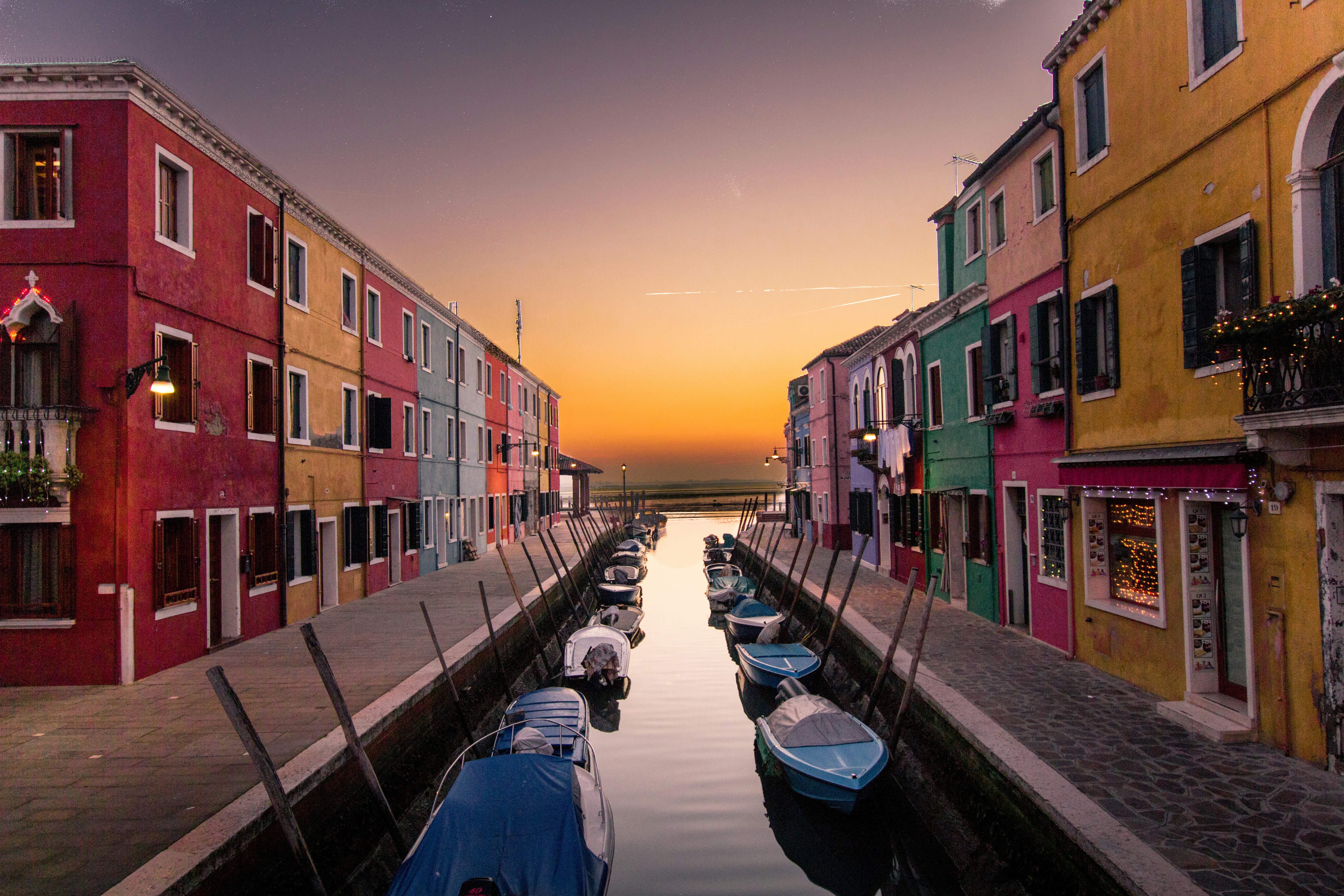 A serene canal scene at sunset with vibrant, colorful houses lining both sides of the waterway. Small boats are moored along the canal, reflecting in the calm water under a golden-orange sky.