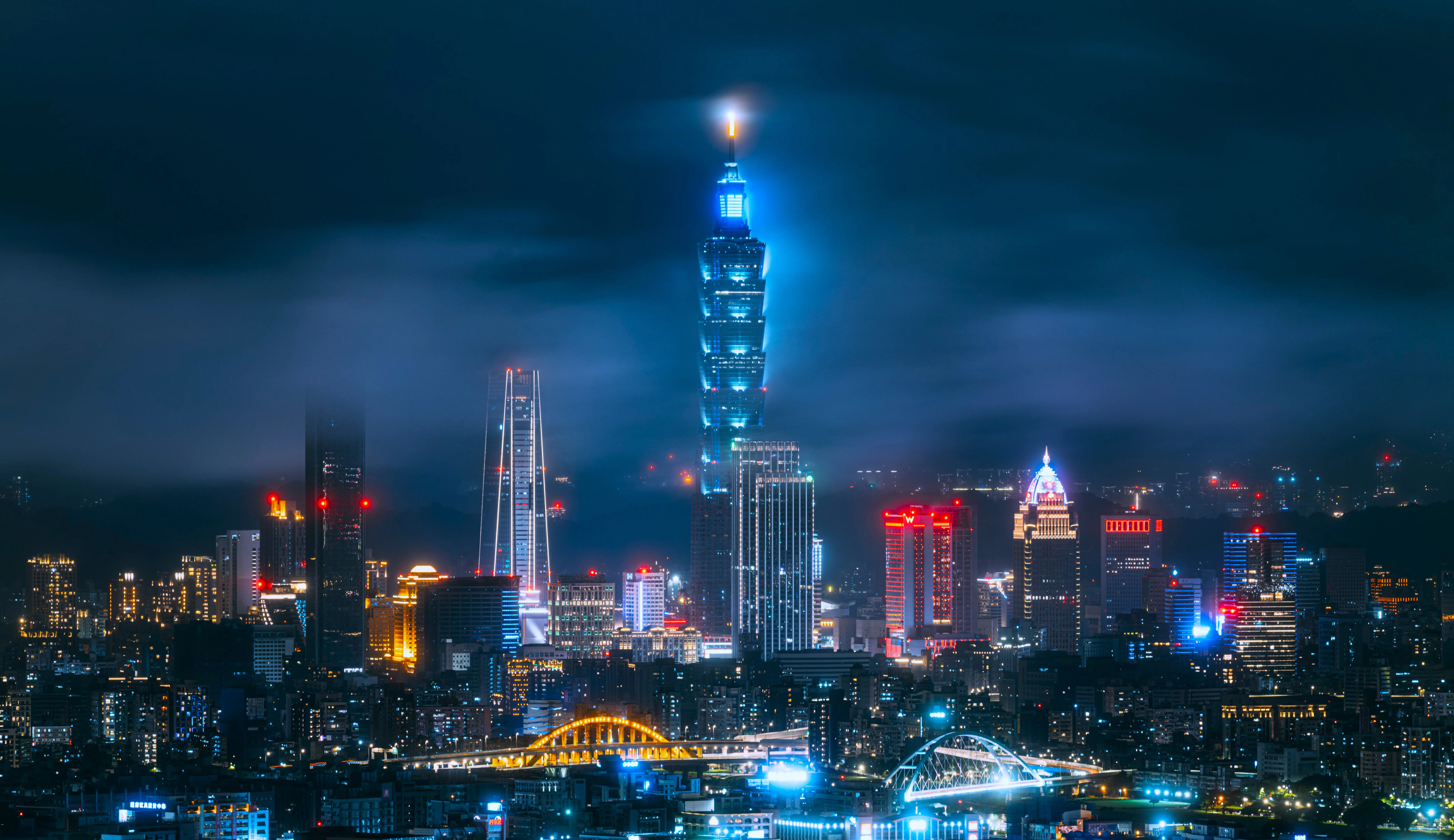 Nighttime skyline of Taipei featuring the illuminated Taipei 101 skyscraper surrounded by modern high-rise buildings and city lights